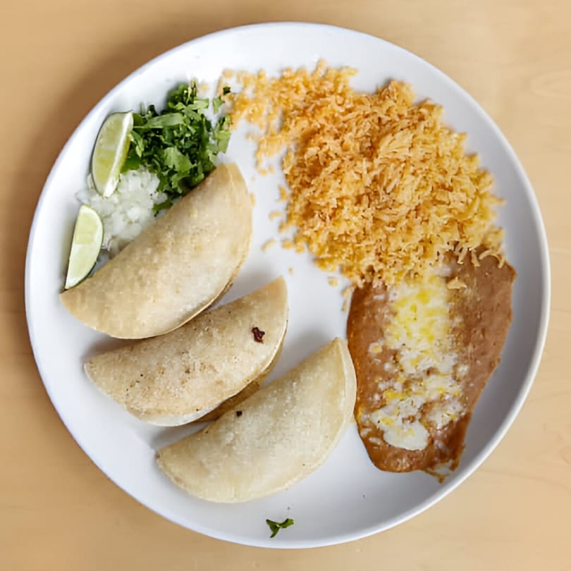 Three empanadas, rice, refried beans, cilantro, onions, and lime wedges on a white plate.