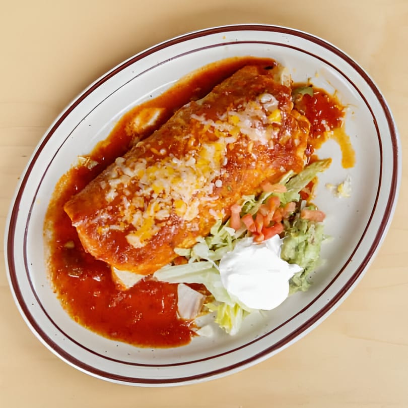 A large, saucy burrito covered in melted cheese, served with guacamole, sour cream, and lettuce.
